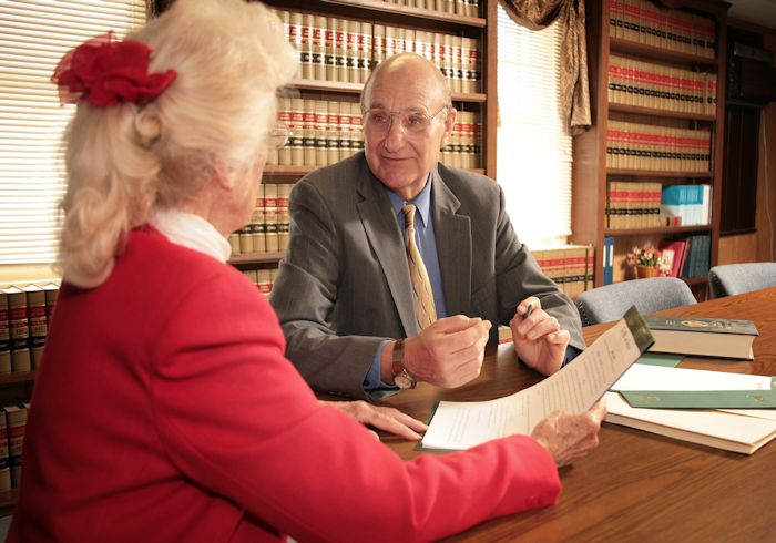 A layer speaks with an elderly client in a red shirt over a piece of paper in a large library with many volumes of law text